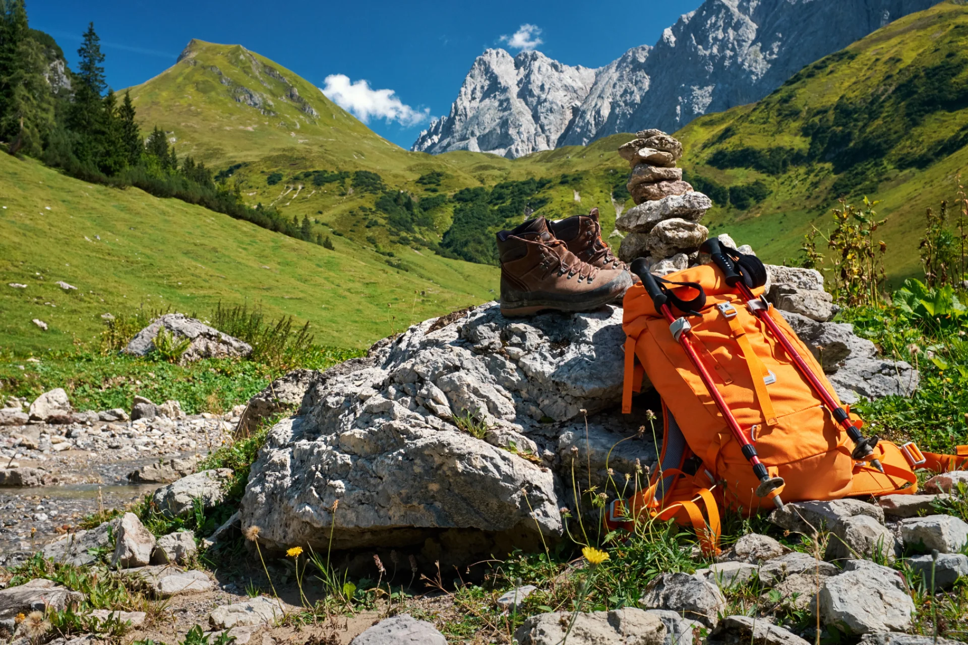 Hiking boots, an orange backpack, and trekking poles resting on rocks in a sunny alpine meadow with tall, rugged mountains in the background.